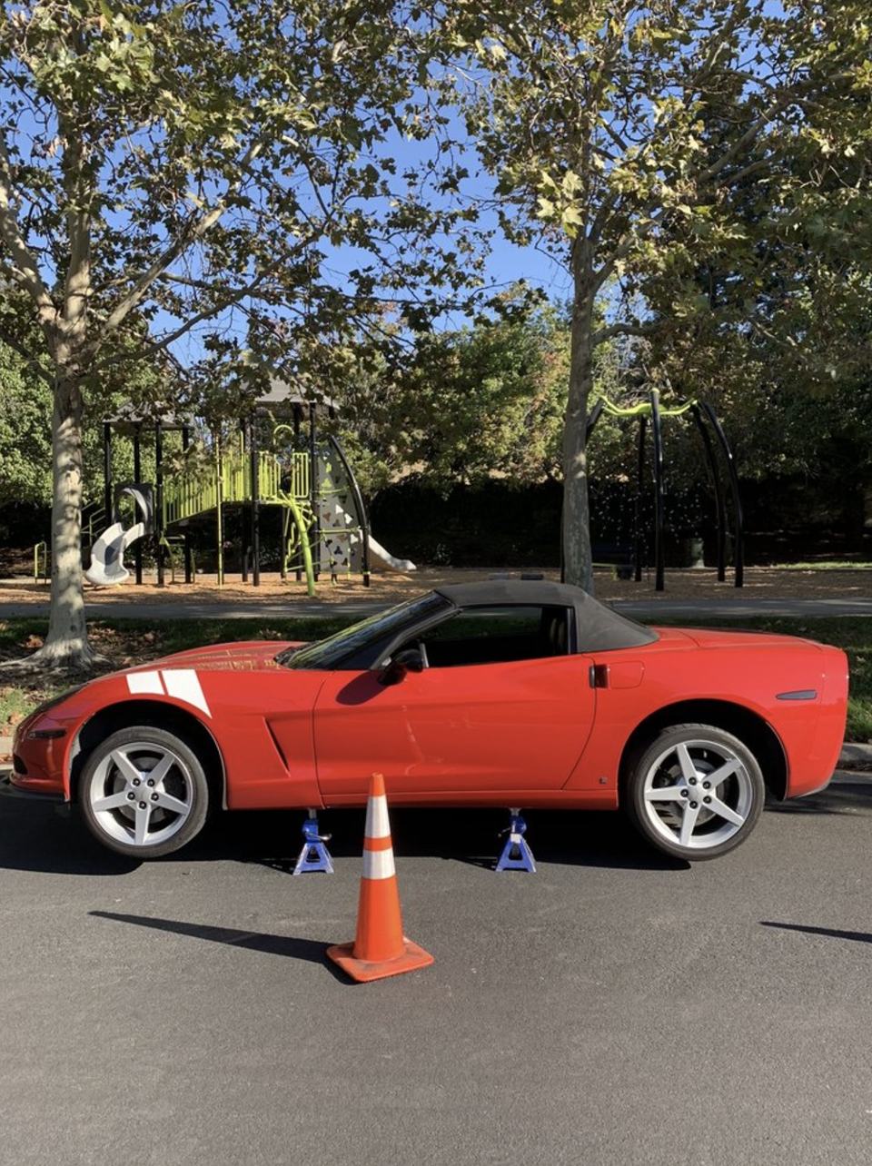 A red Corvette convertible up on jack stands in a parking lot, with an orange cone set out beside it