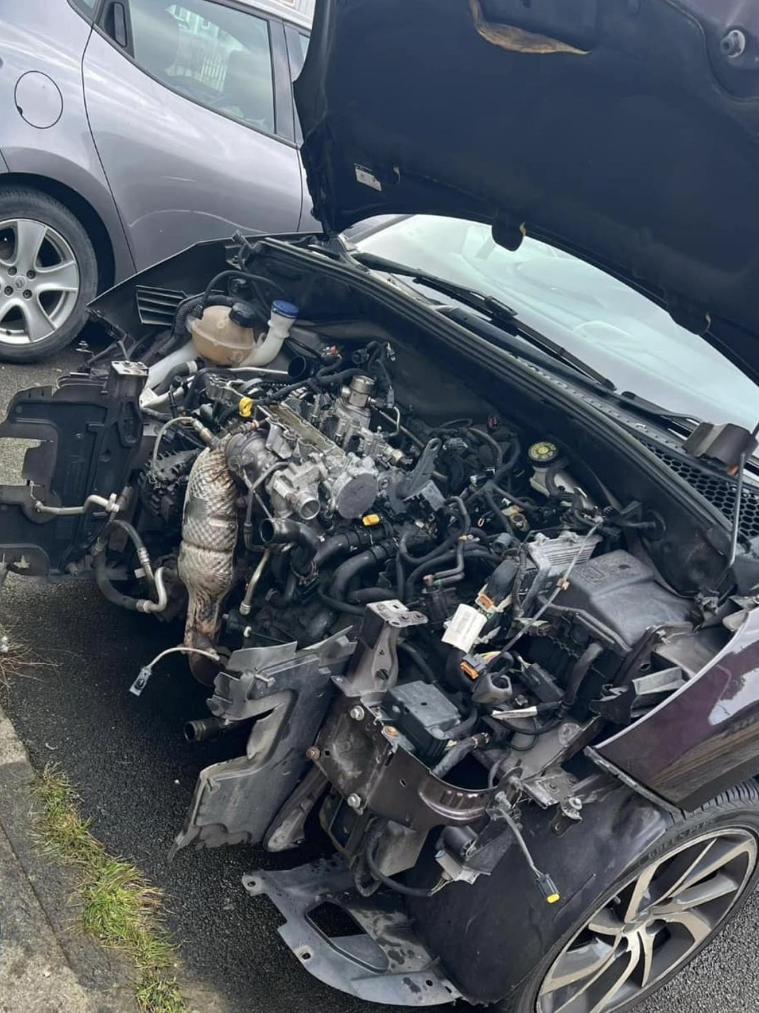 A car with its front bumper and headlights removed, engine exposed for repair at the curb
