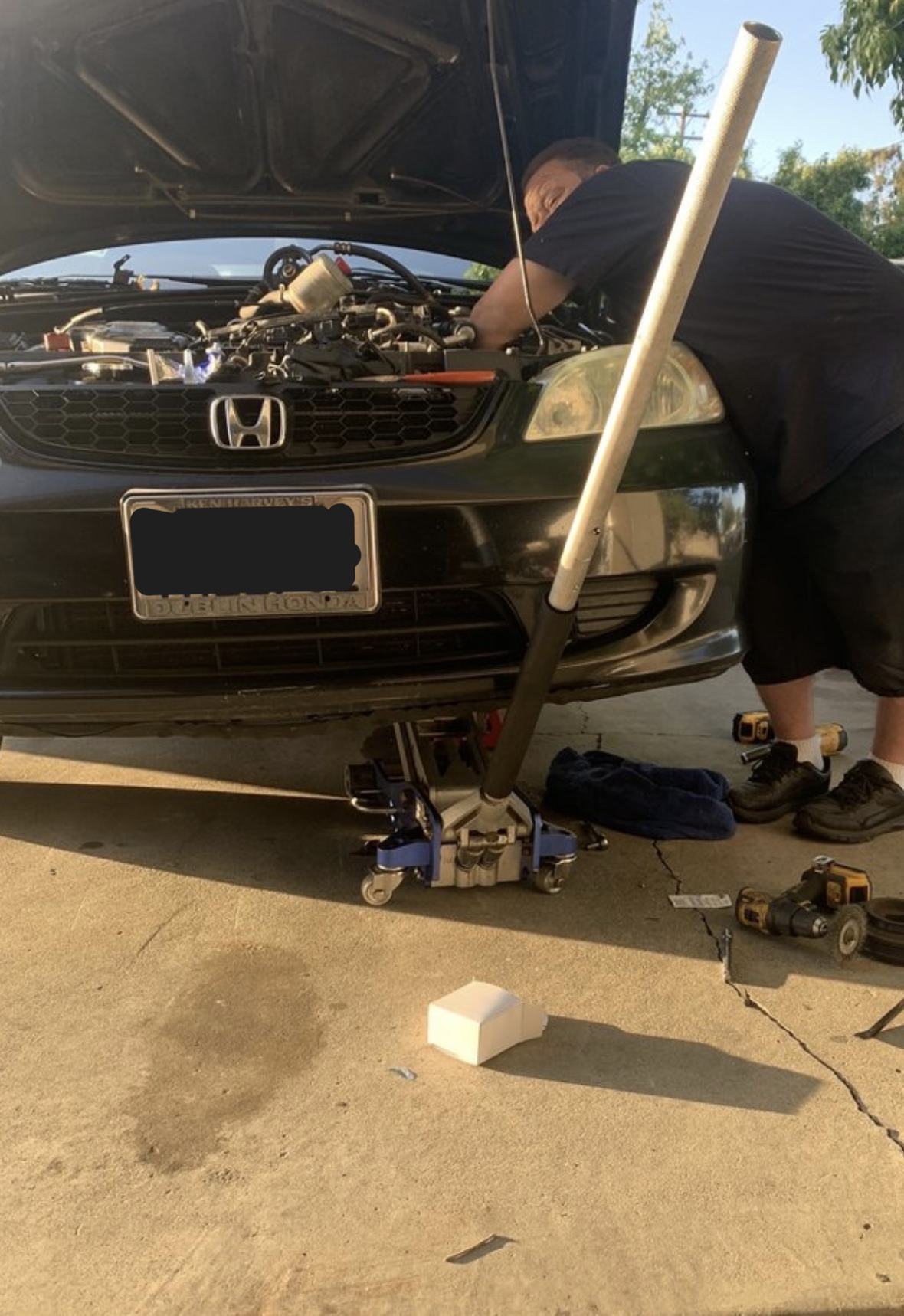A mechanic leaning into the engine bay of a black Honda on a floor jack in a driveway